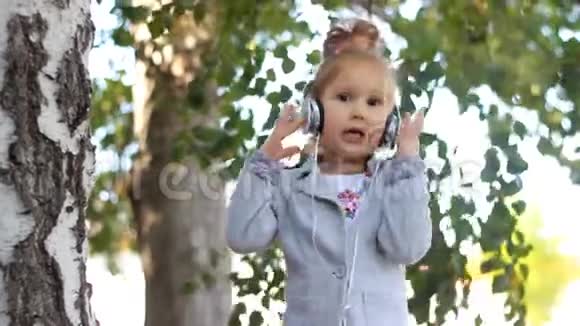 可爱的女孩戴着耳机听音乐在公园里用桦树唱歌视频的预览图
