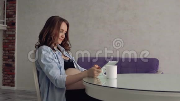 幸福的孕妇坐在玻璃餐桌旁喝咖啡用平板电脑在线讲座视频的预览图