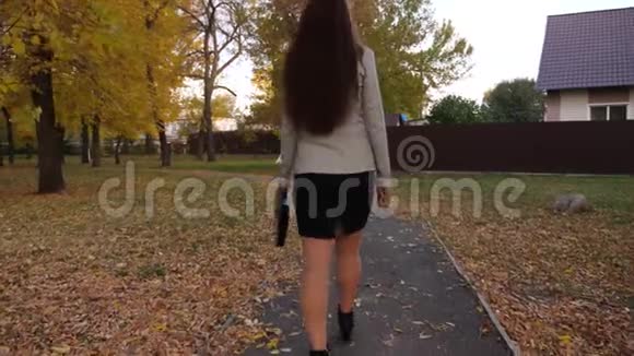 女商人穿着裙子和夹克手里拿着黑色公文包沿着人行道回家穿过秋天的公园视频的预览图