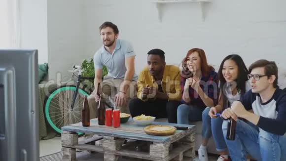 多民族朋友体育迷在电视上观看体育赛事一起在家吃零食喝啤酒视频的预览图
