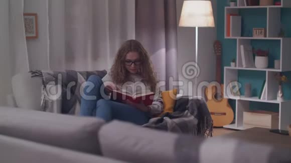 年轻的美女晚上在她舒适的客厅里读一本纸书视频的预览图