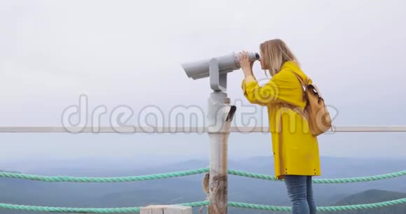 穿着黄色雨衣的女人透过观光望远镜看山视频的预览图