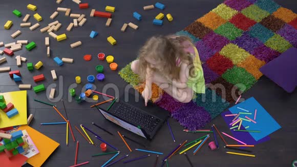 金发女孩子在笔记本电脑上看卡通坐在家里的地板上视频的预览图
