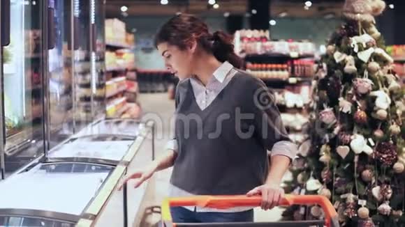 年轻的黑发女孩从冰箱里拿了一包黄油在当地杂货店购物视频的预览图