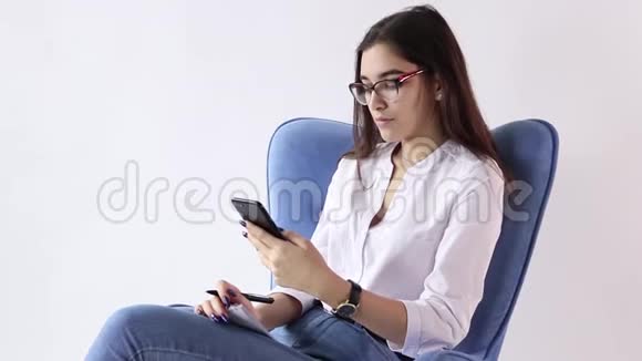 微笑的黑发女人坐在扶手椅上在白色背景下使用智能手机视频的预览图