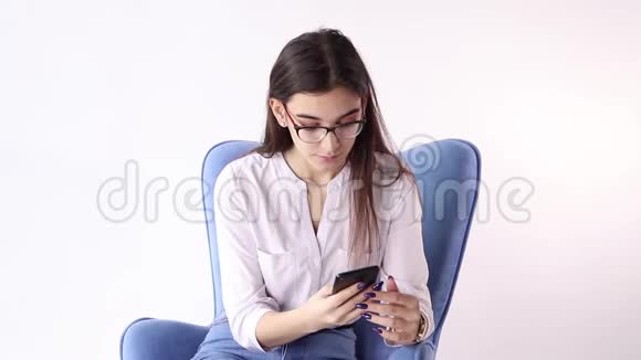 微笑的黑发女人坐在扶手椅上在白色背景下使用智能手机视频的预览图