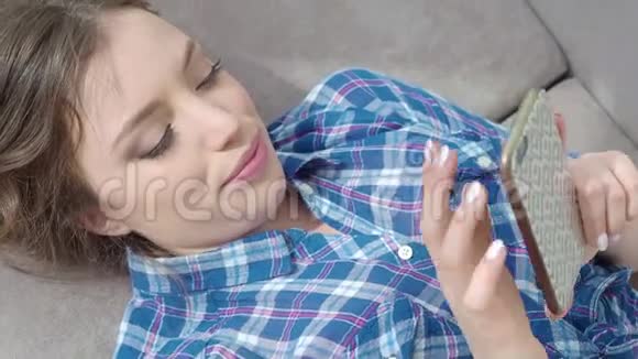 漂亮的年轻女人躺在沙发上在电话里发短信年轻女子用手机微笑着视频的预览图