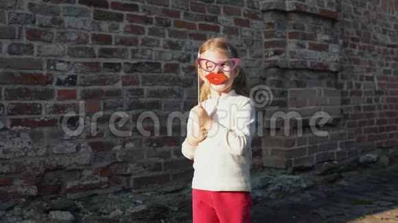 小女孩把纸唇放在砖墙旁边的棍子上视频的预览图