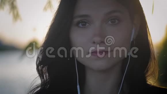 日落背景下戴着耳机的年轻漂亮黑发女人的肖像视频的预览图