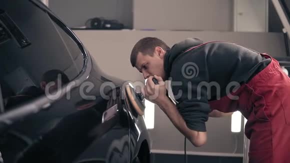 汽车中心的年轻男工人照看着一辆黑色昂贵的汽车用磨砂机抛光擦亮眼睛视频的预览图