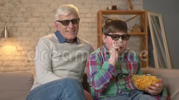 祖父和孙子坐在沙发上戴着3D眼镜看一部3D电影吃着薯片笑着笑着视频的预览图