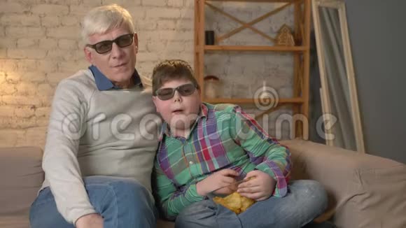 祖父和孙子坐在沙发上看着3D眼镜的3D电影吃薯片搬家电视表演视频的预览图