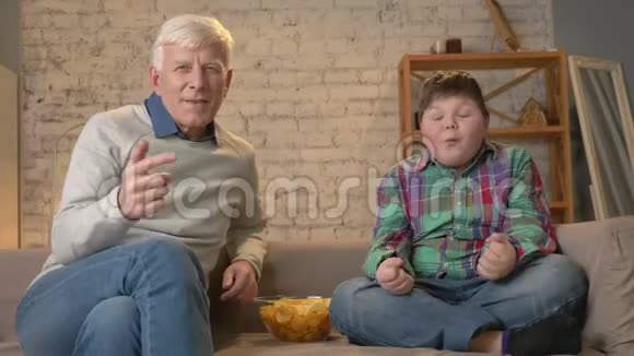 祖父和孙子坐在沙发上看电视吃薯片享受胜利粉丝快乐微笑视频的预览图