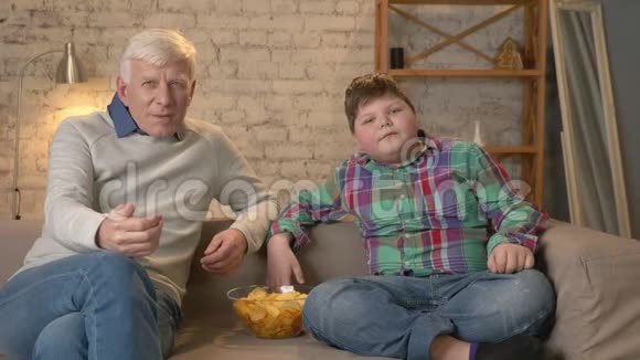 祖父和孙子坐在沙发上看电视吃薯片在胜利中欢欣鼓舞球迷家庭视频的预览图