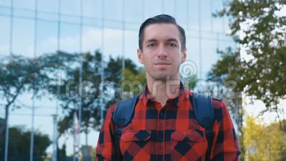 穿格子衬衫和背包看相机的年轻人的肖像大玻璃城市建筑背景视频的预览图