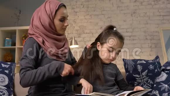 一位年轻的印度母亲戴着头巾给她的小女儿做发型而她的孩子则坐在上面看书视频的预览图