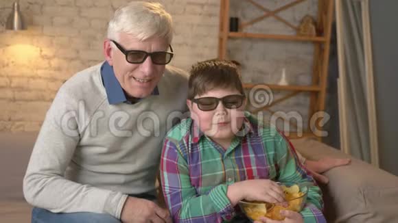 祖父和孙子坐在沙发上看3D眼镜片吃薯片说话跳舞视频的预览图