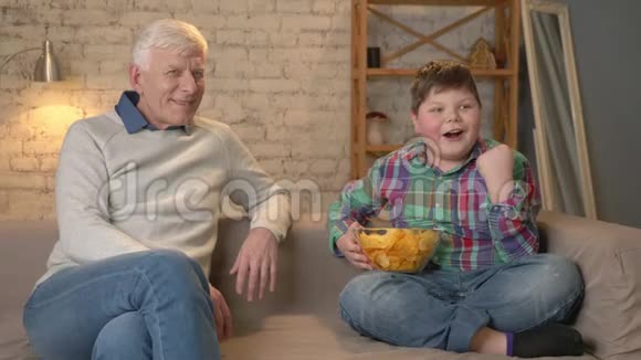 爷爷和孙子坐在沙发上看电视胖男孩给爷爷吃薯片视频的预览图