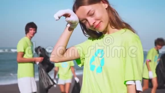 四名志愿者穿着绿色T恤衫带着图像回收在海滩上收集垃圾拿着收集的袋子在镜头前看视频的预览图