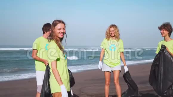 四名志愿者穿着绿色T恤衫带着图像回收在海滩上收集垃圾拿着收集的袋子在镜头前看视频的预览图