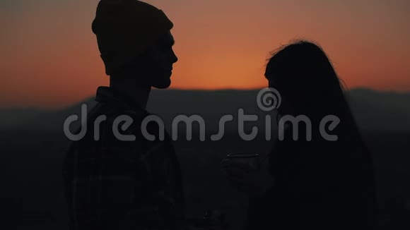 在美丽的夕阳背景下一对相爱的年轻夫妇用热水瓶喝茶的剪影视频的预览图