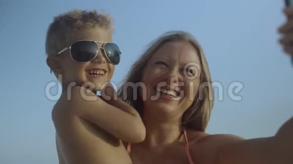 年轻漂亮积极的母亲正和她的小儿子坐在一起在海滩上的智能手机上自拍视频的预览图