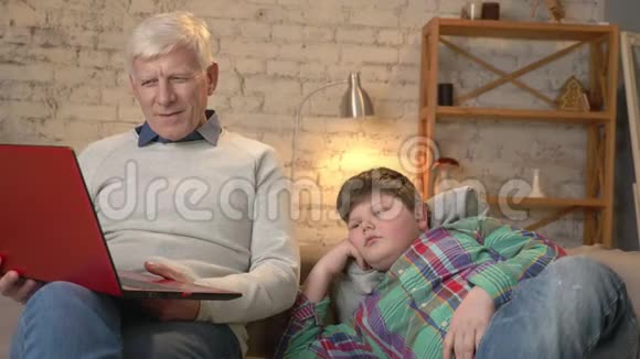 祖父和孙子坐在沙发上在笔记本电脑上看着一段有趣的视频小胖子是视频的预览图