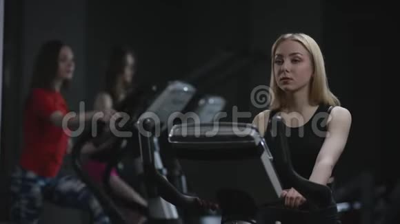 漂亮的金发女孩正在健身车上辛勤工作酷男在练习的背景下打招呼视频的预览图