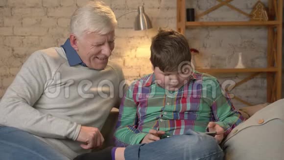 一个年轻的胖男孩和他的祖父坐在沙发上玩平板电脑孙子教爷爷视频的预览图