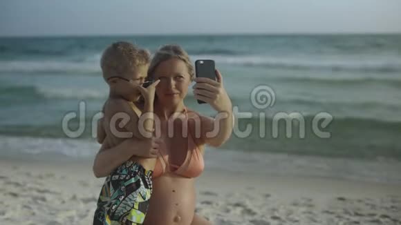 年轻漂亮积极的母亲正和她的小儿子坐在一起在海滩上的智能手机上自拍视频的预览图