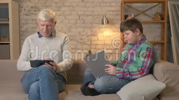 代的差异一位老人正在用平板电脑和耳机坐在沙发上一个年轻的胖男孩正在视频的预览图
