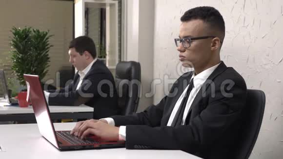 年轻成功的非洲商人戴着眼镜坐在办公室里用笔记本电脑工作脱下他的眼镜视频的预览图