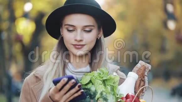 快乐的年轻美丽的女人穿着时尚的外套在公园里散步拿着一包产品用智能手机视频的预览图