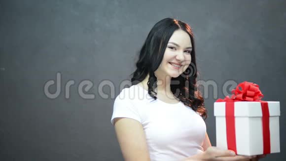 年轻的黑发女人在灰色背景下送礼物带红色丝带的白色礼品盒视频的预览图