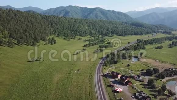 阿尔泰山上一条铺着沥青的蜿蜒公路在一片绿油油的田野里在一条绿树成荫的河流附近的空中俯瞰视频的预览图
