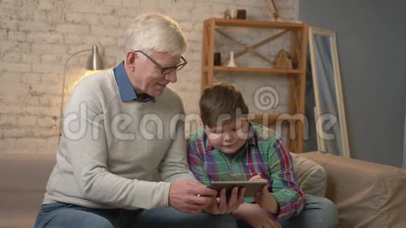 祖父和孙子坐在沙发上用平板互相看着微笑着家庭舒适家庭懒散视频的预览图