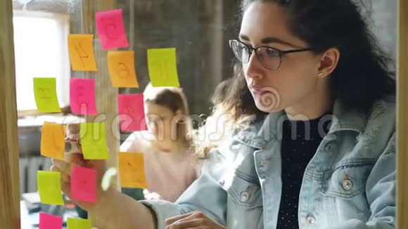 年轻有创意的女商人在与女性合作的同时在玻璃板上编号彩色贴纸视频的预览图