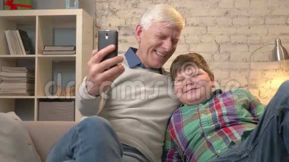 祖父和孙子坐在沙发上老人手里拿着智能手机自拍他们视频的预览图
