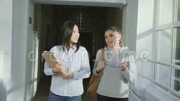 两个年轻漂亮的女学生走在宽阔而轻盈的大学走廊里积极地微笑着讨论家庭作业视频的预览图