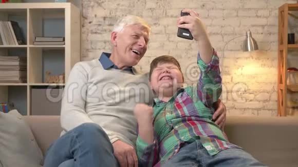 爷爷和孙子坐在沙发上用智能手机自拍在胜利中欢乐他们是视频的预览图