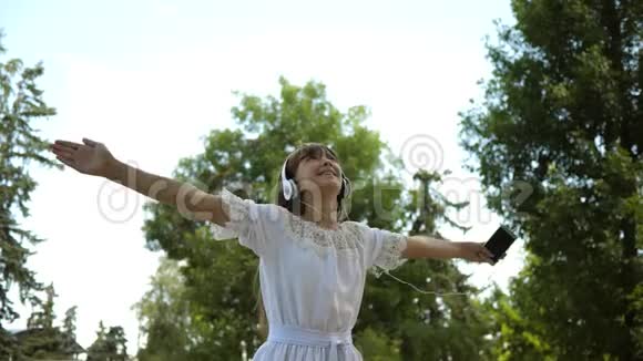 快乐女孩戴着耳机微笑手里拿着智能手机在夏天的公园里跳舞慢动作美丽的姑娘视频的预览图