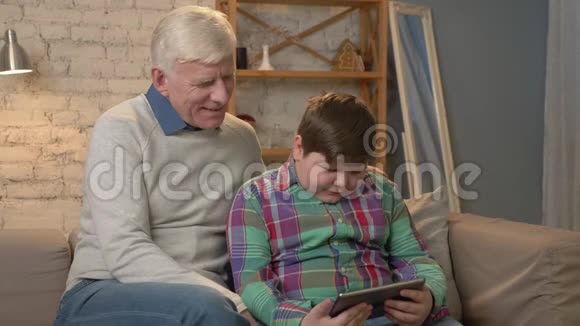 祖父和孙子坐在沙发上用平板电脑看着相机家庭舒适家庭懒散视频的预览图