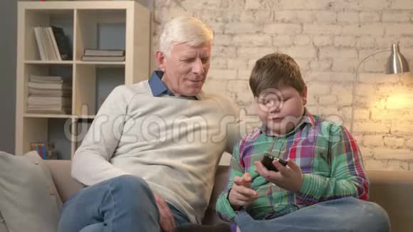 爷爷和孙子坐在沙发上用智能手机做自拍小胖孩子和爷爷男孩视频的预览图
