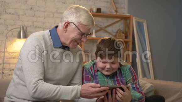 祖父和孙子坐在沙发上用平板电脑看着一部有趣的电影笑着微笑着家庭舒适视频的预览图