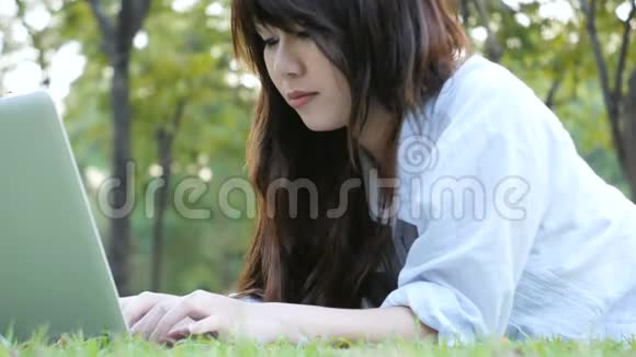 年轻的亚洲女人用开着笔记本电脑在绿草上双腿女孩手在键盘上远程学习概念视频的预览图