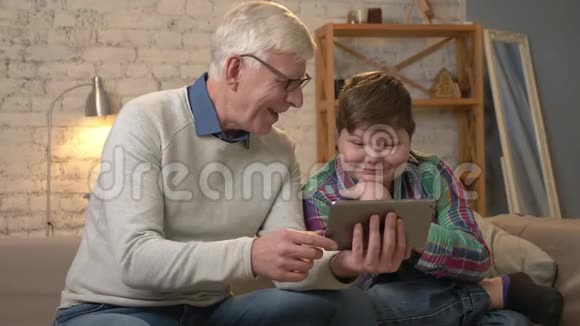 爷爷和孙子正坐在沙发上用平板电脑挥手在视频中聊天打招呼微笑视频的预览图