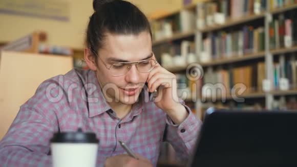 坐在大学图书馆里用智能手机聊天的年轻帅哥戴眼镜男学生的特写镜头视频的预览图