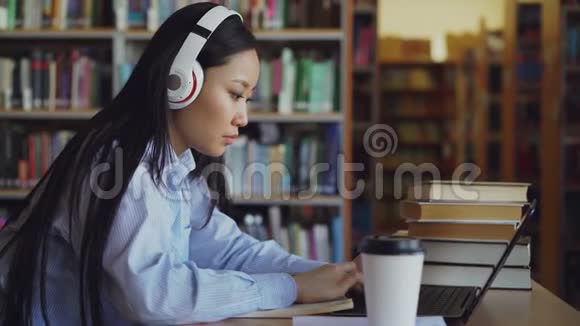 漂亮的亚洲女学生戴着耳机坐在图书馆的办公桌前工作集中在学校项目上青年视频的预览图
