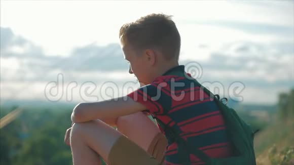 一个年轻的白种人少年穿着一件红色衬衫背着一个背包日落时分坐在高高的山上视频的预览图