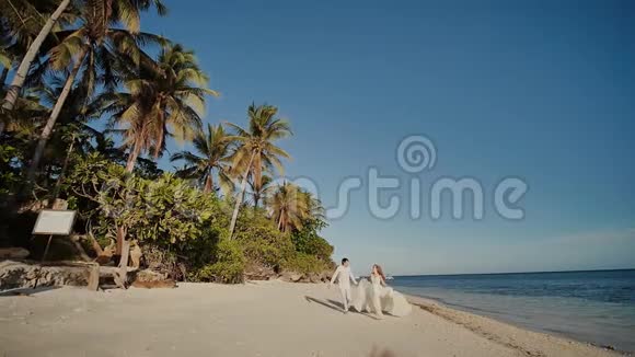新郎和美丽的新娘赤着脚沿着海边的沙滩奔跑一起快乐一场热带婚礼视频的预览图